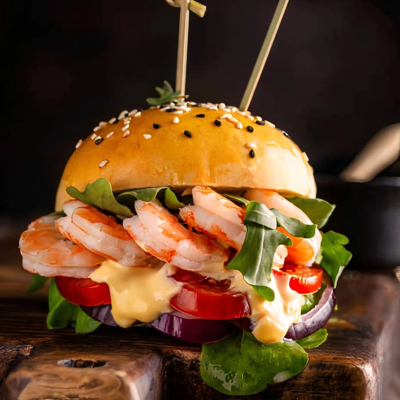 A Dakari Soul Food shrimp burger with lettuce, tomato, onion, and sauce on a wooden board.